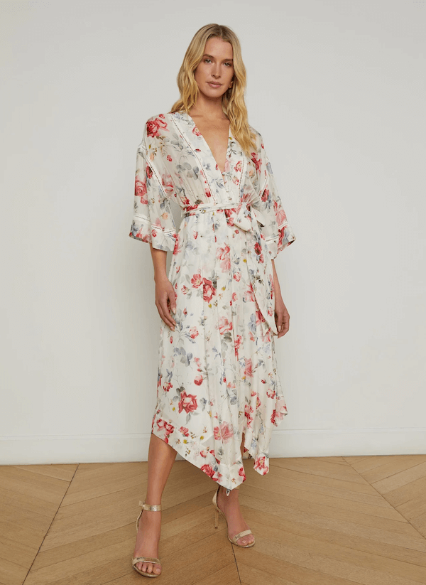 A woman stands indoors after memphis shopping, wearing a white, floral-print, belted wrap dress with wide sleeves and an asymmetrical hem, paired with strappy heels.