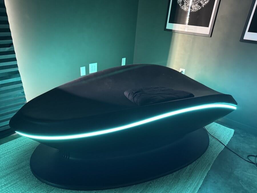 A black, futuristic SAVA Sound Pod lounge chair with a curved design and blue LED strip light sits on a rug in a modern room with framed art, delivering an immersive sound experience.
