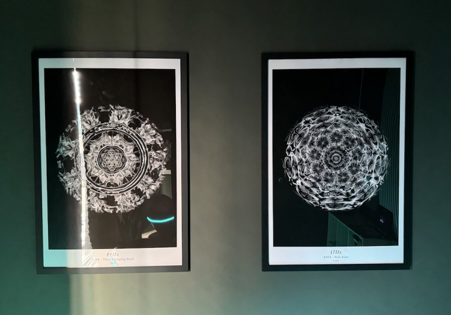Two framed black-and-white geometric art prints hang side by side on a dark wall, with light reflecting off the glass of the left frame inside a modern SAVA Sound Pod.
