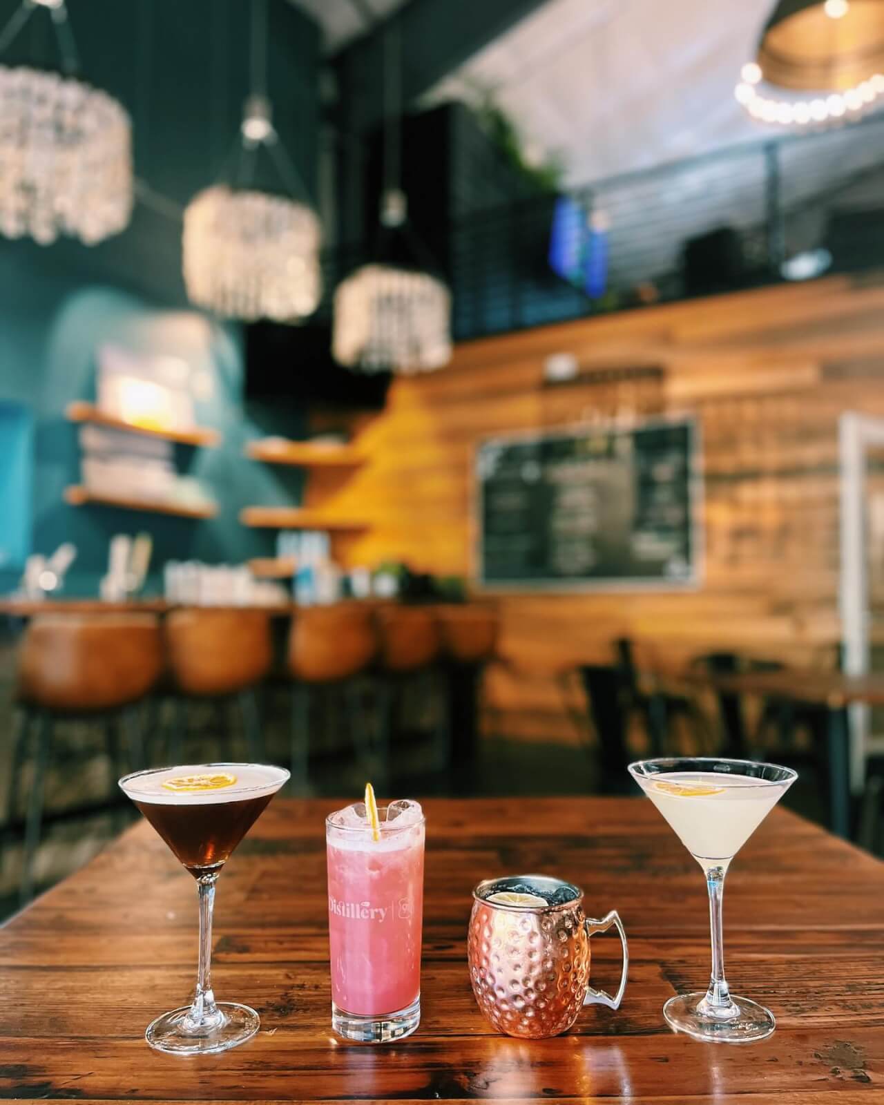 Four assorted cocktails are displayed on a wooden table in a modern 30A bar with teal walls, wooden accents, and hanging light fixtures in the background.