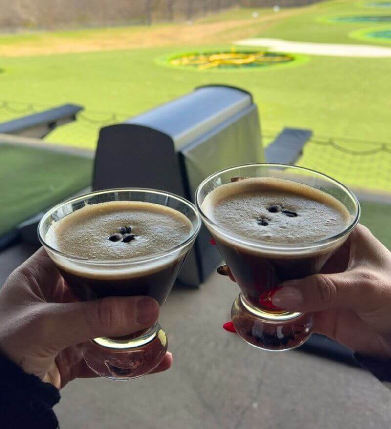 Two hands holding espresso martinis in cocktail glasses, with a Topgolf driving range visible in the background.