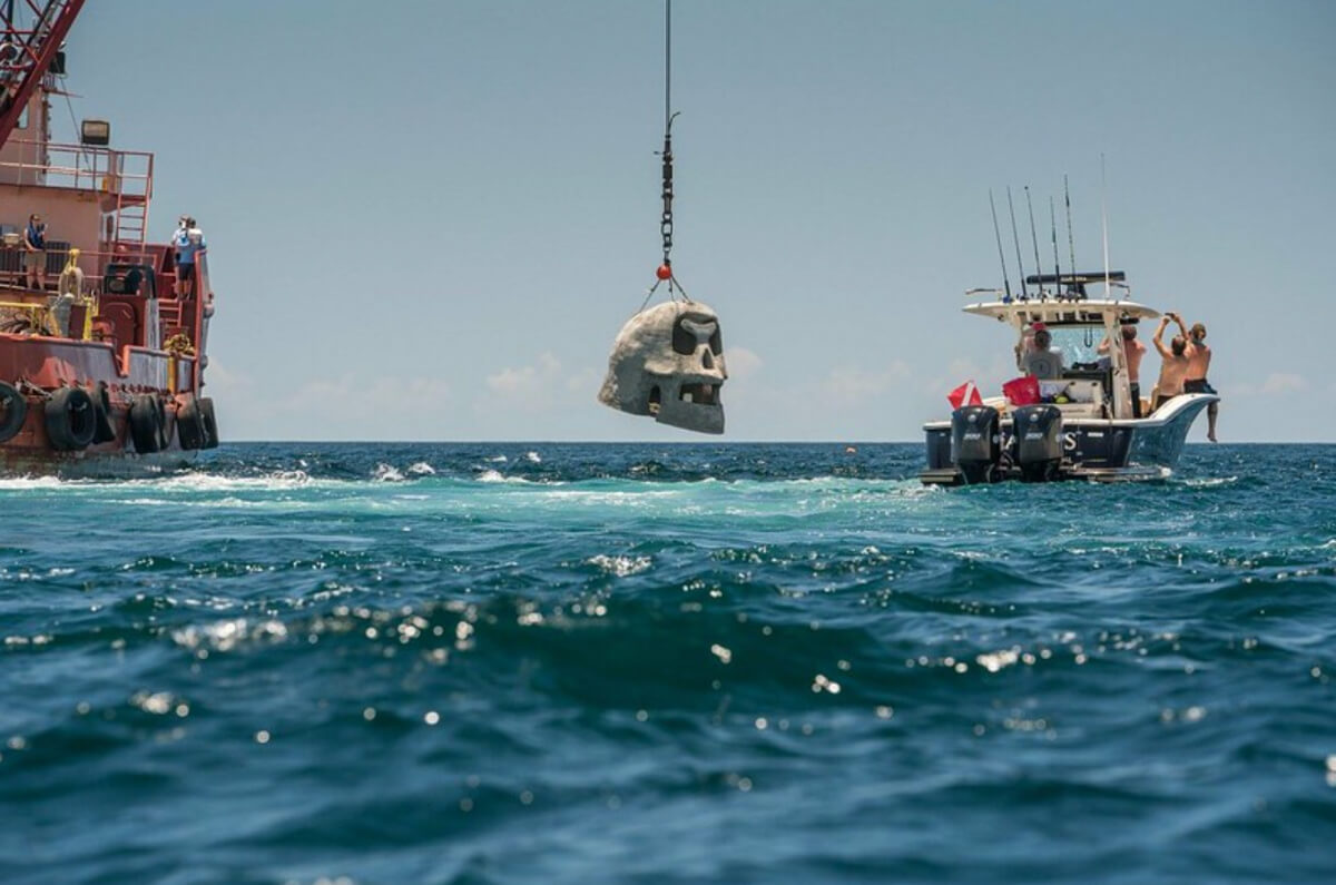 A crane on a barge lowers a large concrete sculpture shaped like a human skull into the ocean near a small fishing boat, as people observe the creation of an underwater museum.