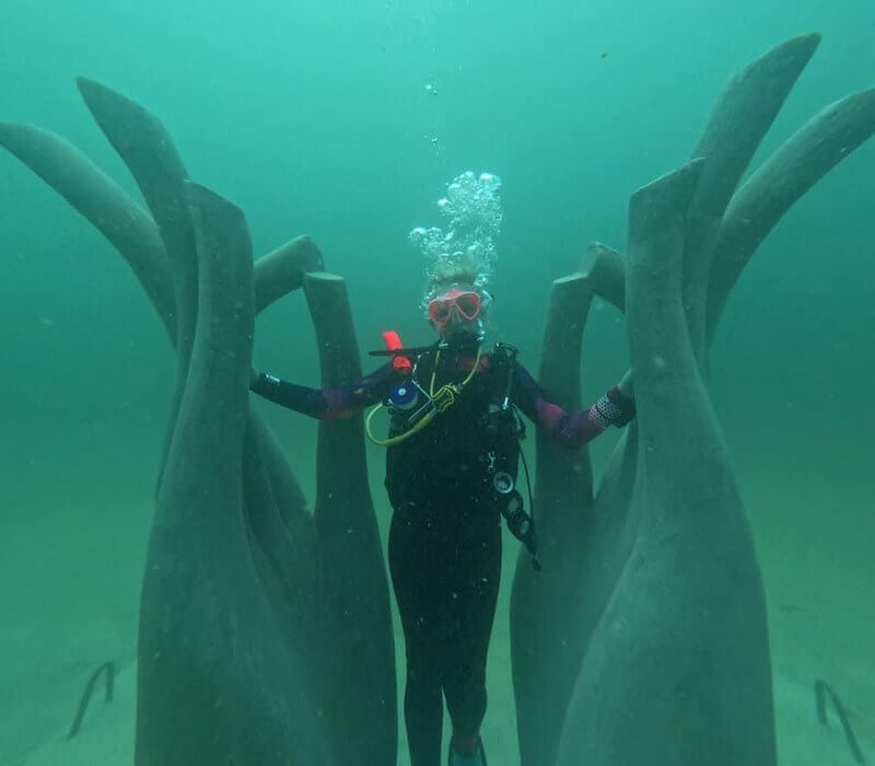 A scuba diver poses underwater between two large, curved, hand-like sculptures on the ocean floor, creating a surreal scene that feels like an underwater museum.