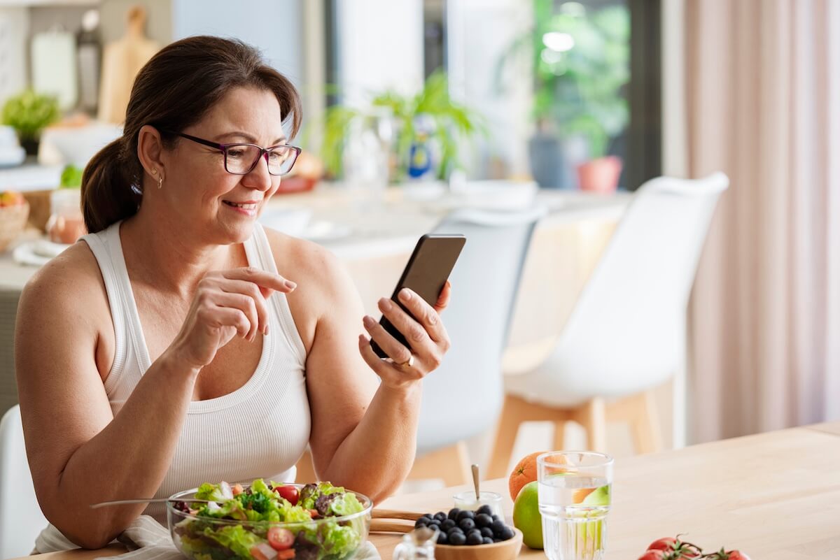 Woman wearing glasses sits at a table with a salad, fruit, and water, smiling while using a wellness app on her smartphone in a bright kitchen.
