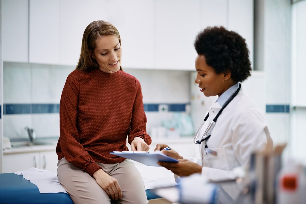 A doctor and a patient sit in a medical office, discussing notes on a clipboard. The patient is smiling and pointing at the clipboard, highlighting the promise of AI in healthcare to enhance patient understanding and care.