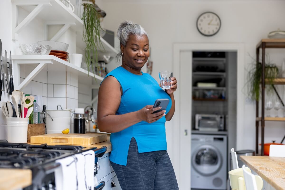 A woman stands in a kitchen, holding a glass of water in one hand and checking her wellness app on her smartphone with the other.