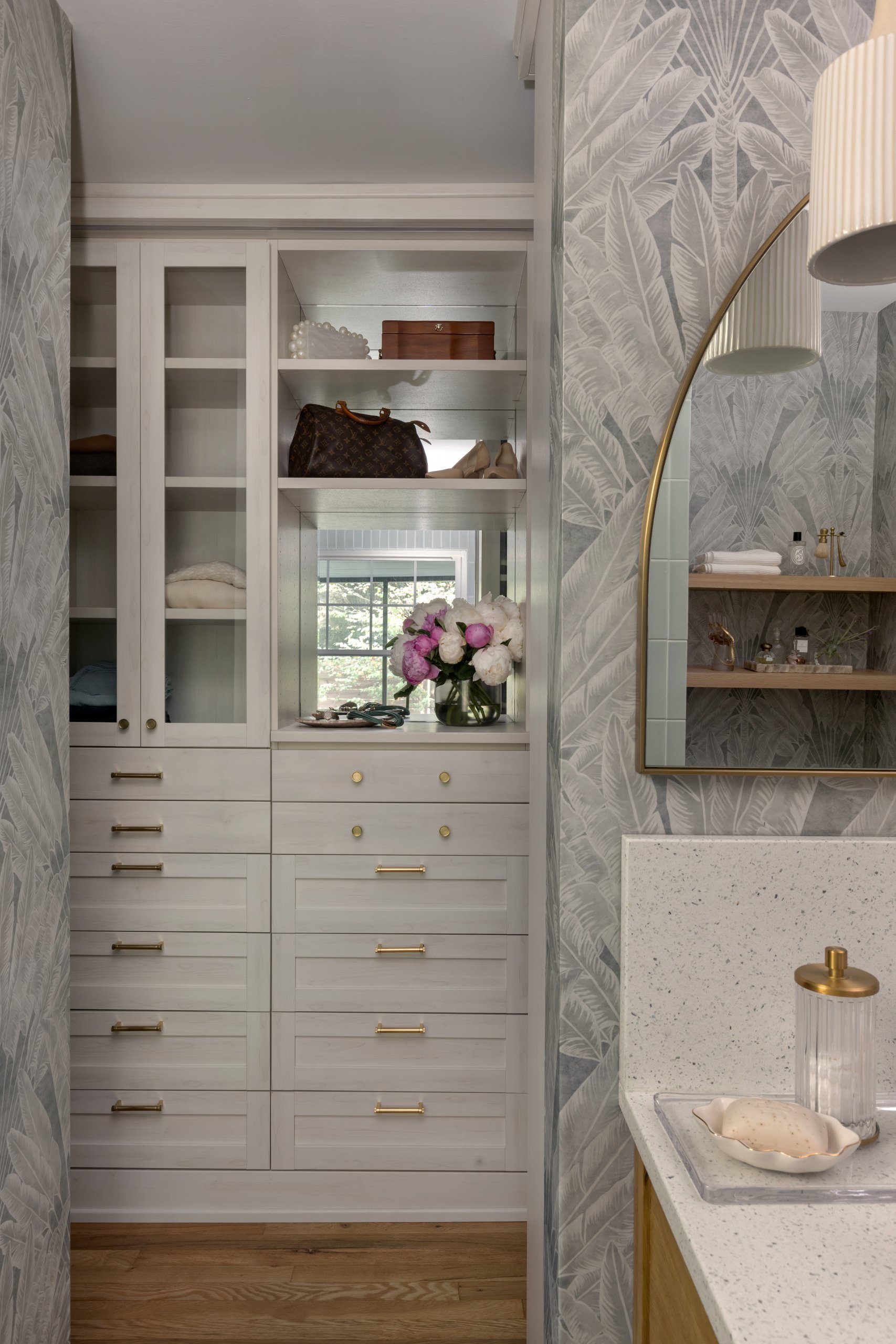 Custom white cabinetry with gold handles, glass-fronted upper shelves, and decorative items including a vase of flowers create a modern, wallpapered dressing area—perfect inspiration for luxury closets and elegant closet design.