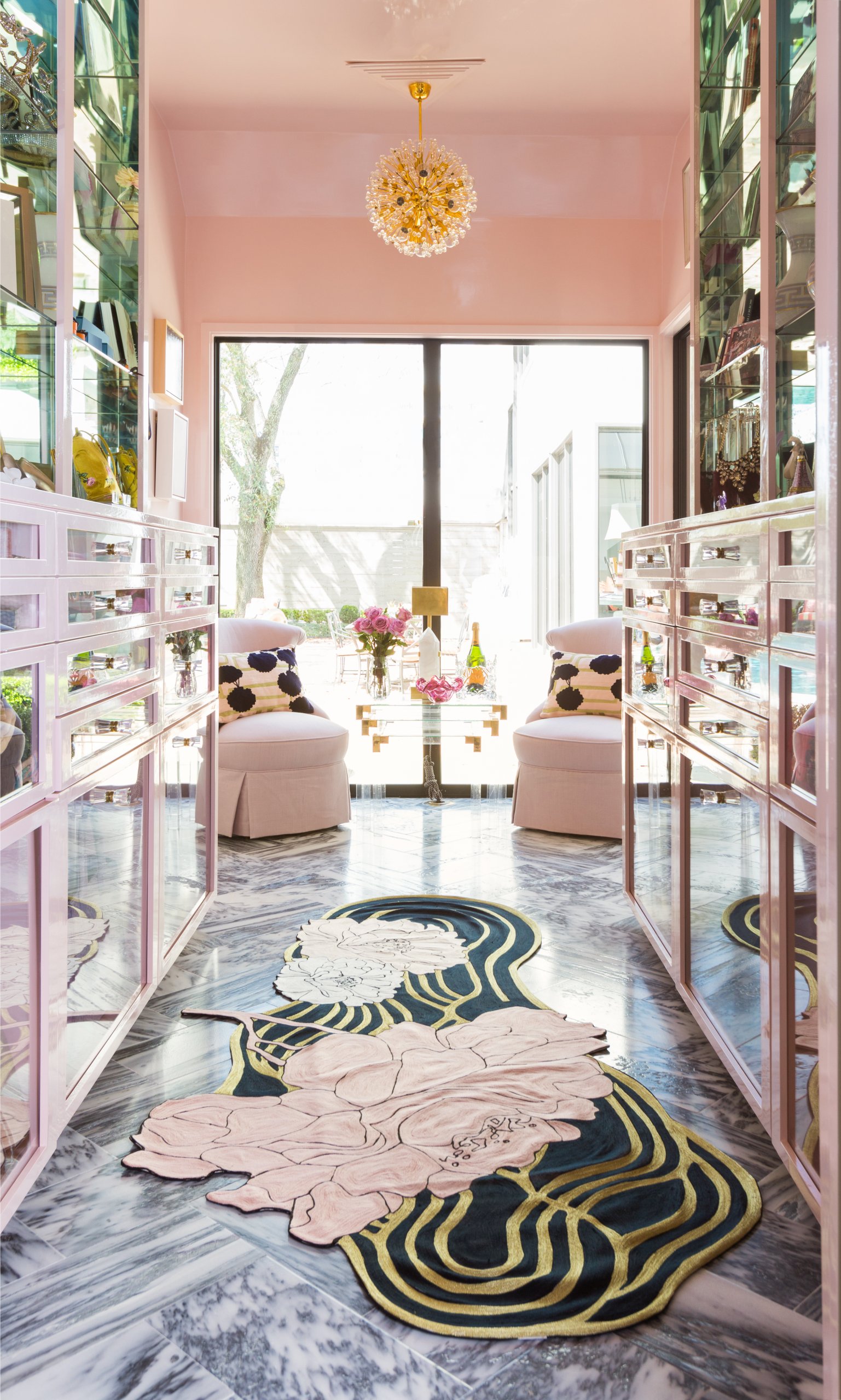 A hallway with pink walls and mirrored cabinets features a modern rug, two round chairs with pillows, and a glass door leading to an outdoor patio—perfect inspiration for luxury closets or elevated closet design.