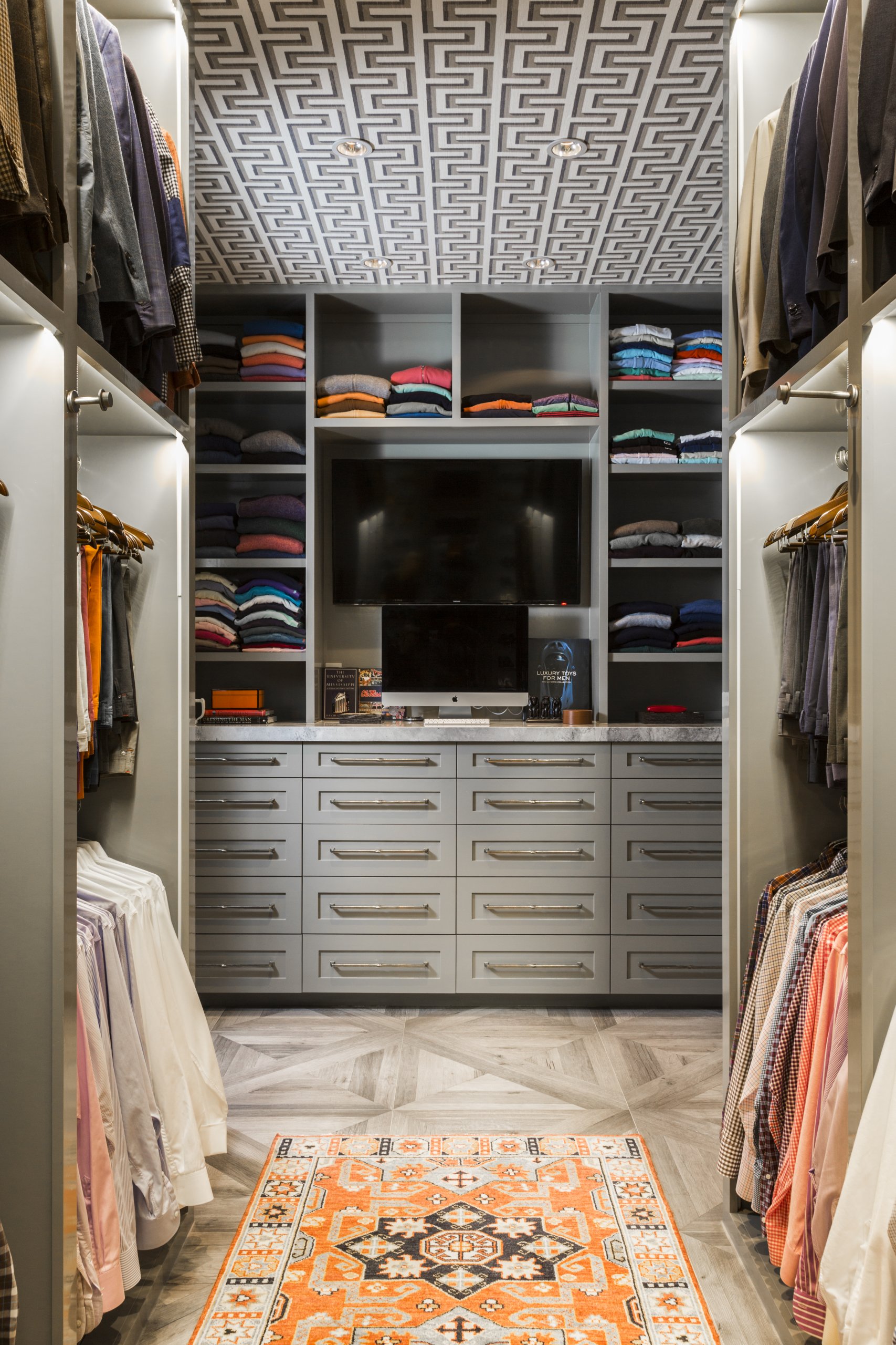A modern walk-in closet featuring smart closet organization with shelves and drawers, a TV on the center dresser, and an orange patterned rug on a herringbone floor.