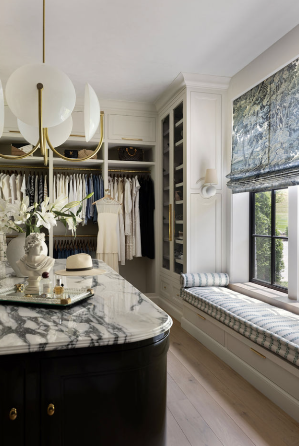 A well-organized walk-in closet with hanging clothes, built-in shelves, a marble-topped island, and a window seat with patterned cushions and a Roman shade showcases exceptional closet design.