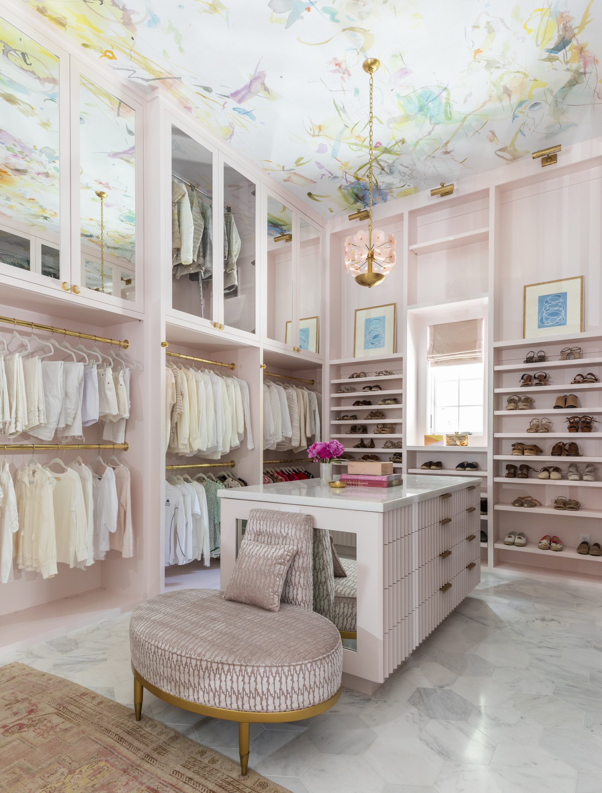 A spacious, pastel pink walk-in closet showcases clever closet organization with clothing racks, shoe shelves, a central island with drawers, a plush chair, floral ceiling, and a pendant light for dreamy closet ideas.