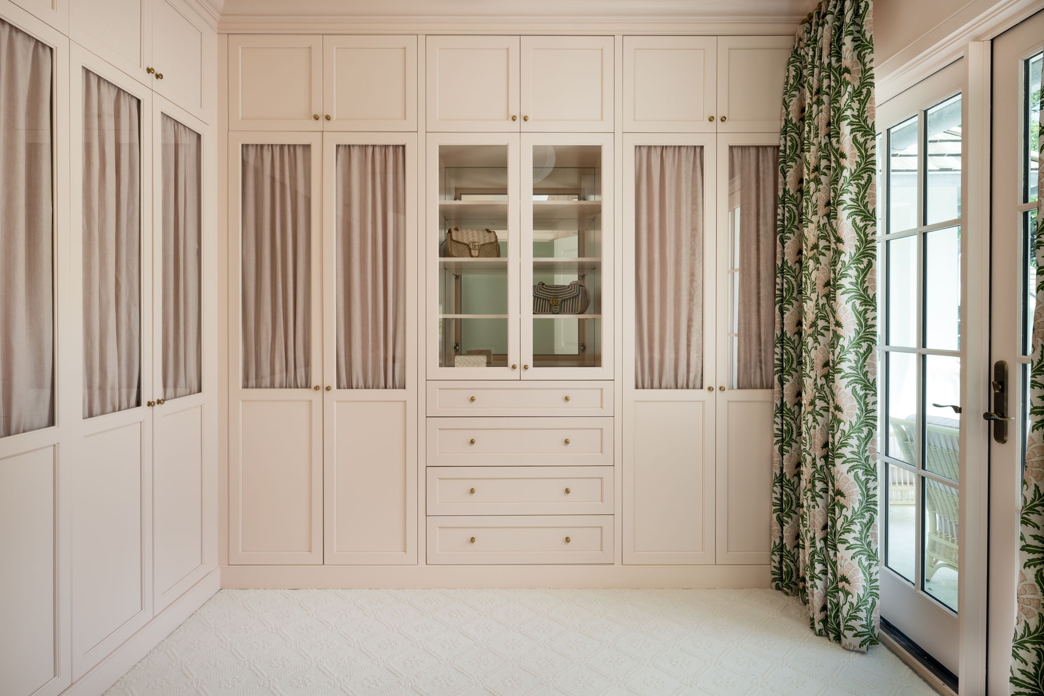 Built-in cream-colored cabinets with glass and fabric panel doors, central drawers, and patterned curtains beside a glass door offer inspiring closet ideas for refined closet design.