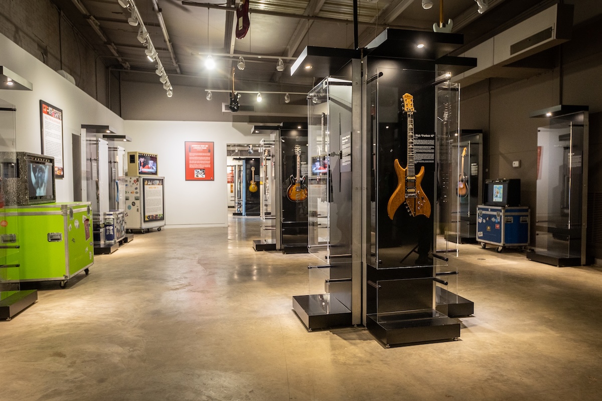 A museum exhibit hall in Greenwood displays various electric guitars in glass cases, with informational plaques and equipment cases placed throughout the room—one of the top things to do in Greenwood for music enthusiasts.