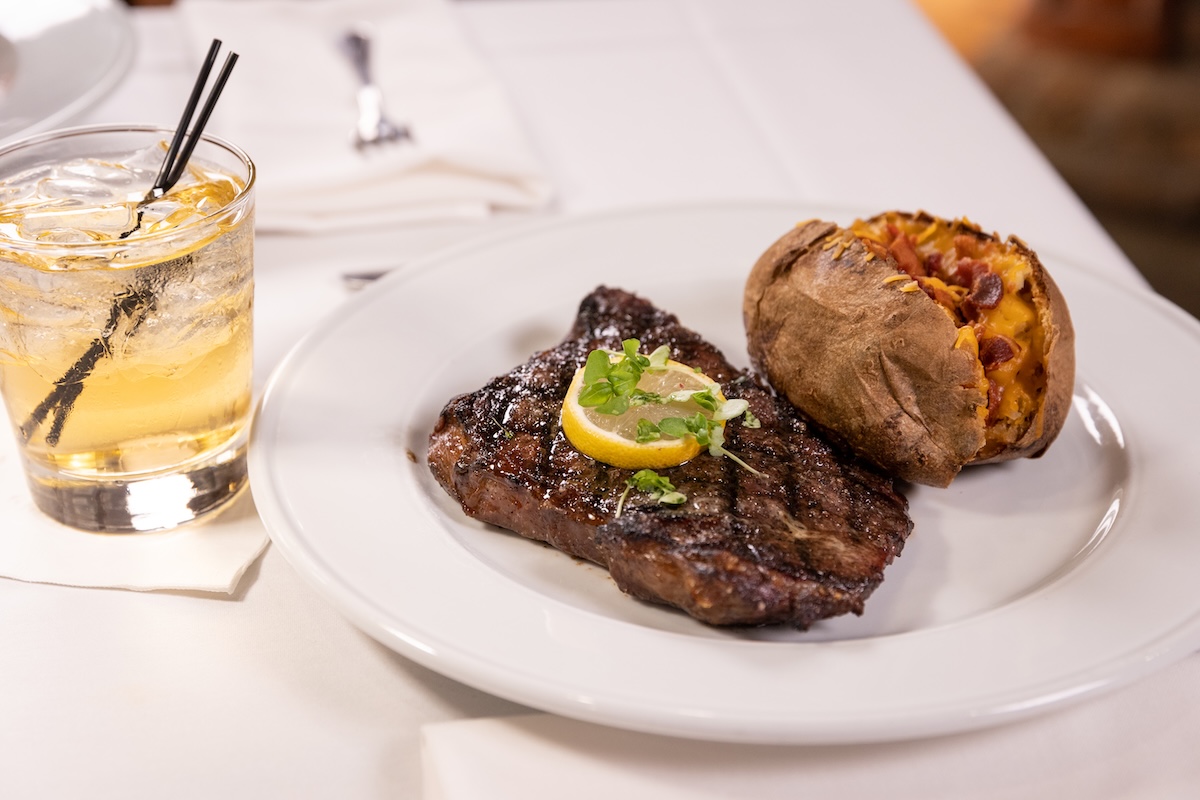 A grilled steak garnished with a lemon slice and herbs is served next to a loaded baked potato on a white plate, perfect for savoring after exploring things to do in Greenwood, with a glass of iced drink beside the plate.
