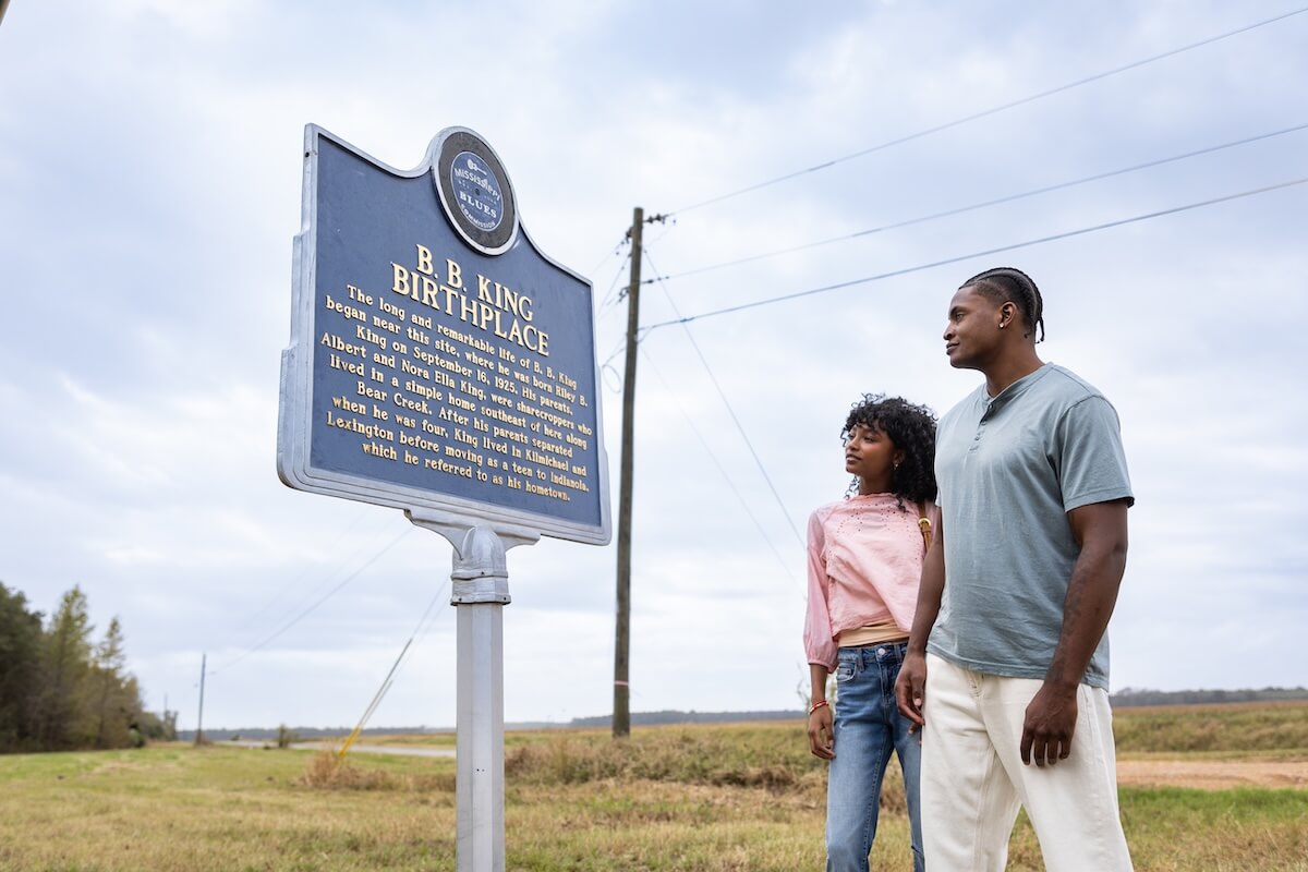 Two people stand beside the B.B. King Birthplace historical marker on a rural roadside, soaking in the local history. Travel to Greenwood MS and visit Greenwood to experience this iconic landmark firsthand.