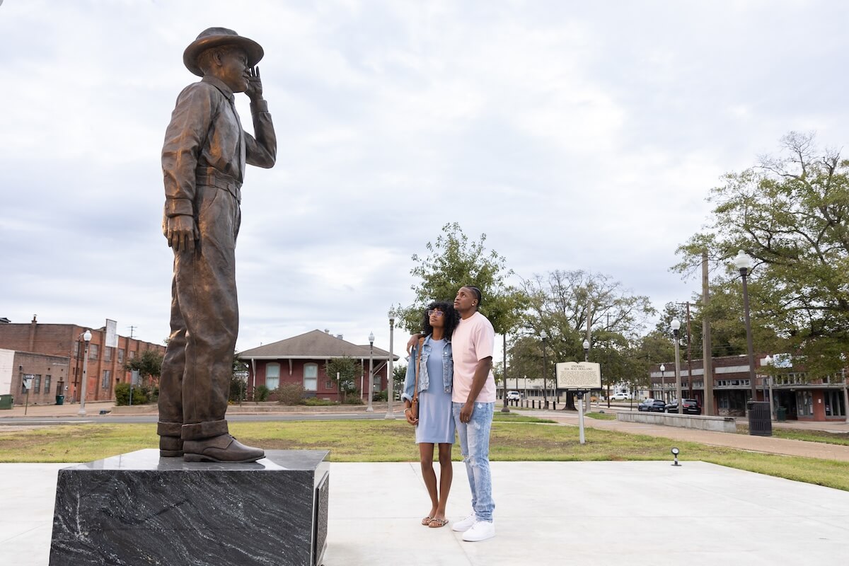 A man and woman stand together outdoors, looking at a large bronze statue of a saluting figure on a pedestal—one of the memorable things to do in Greenwood, with buildings and trees in the background.