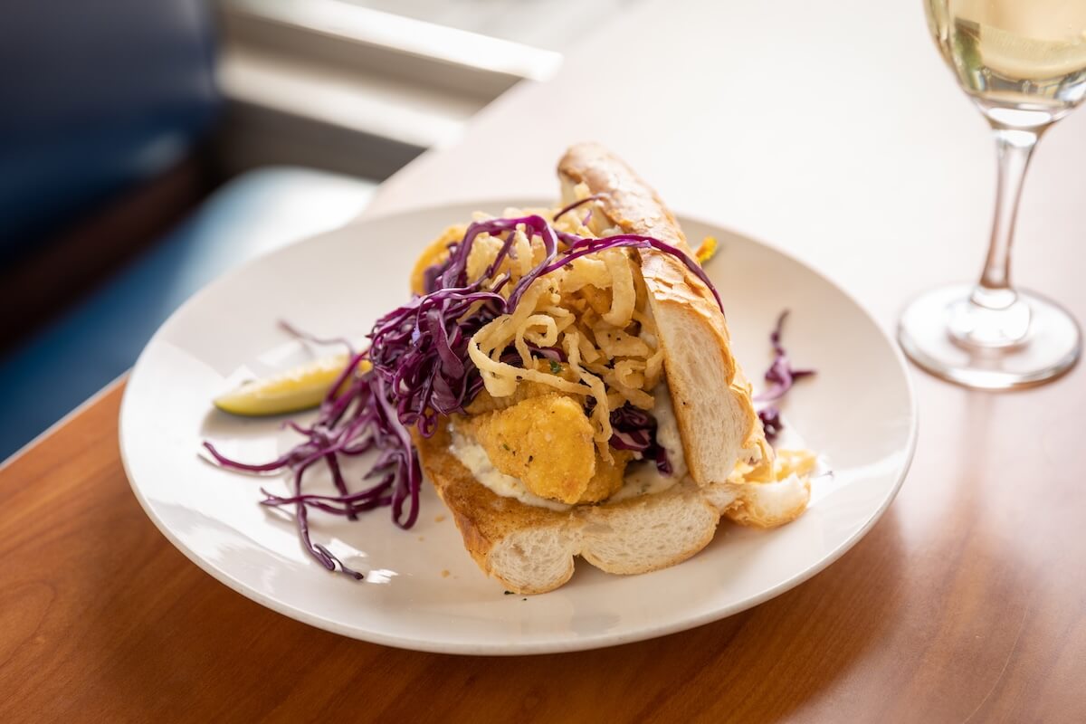 A fish sandwich with fried fish, sliced red cabbage, and fried onions on a hoagie roll, served on a white plate with a lemon wedge and a glass of white wine—one of many tasty reasons to visit Greenwood MS.