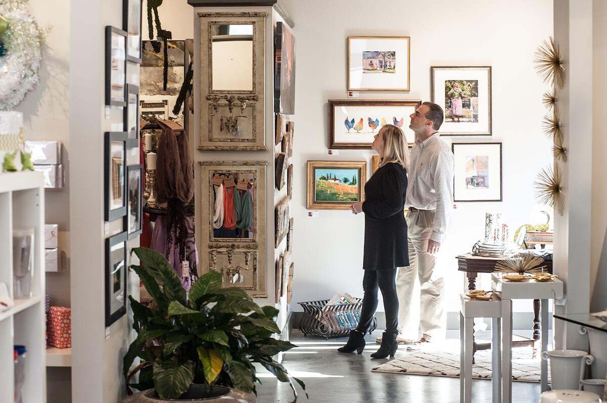A woman and a man stand in an art gallery or shop in Greenwood MS, admiring framed paintings on the wall. The bright space showcases unique art and home items—perfect for those looking for things to do in Greenwood.