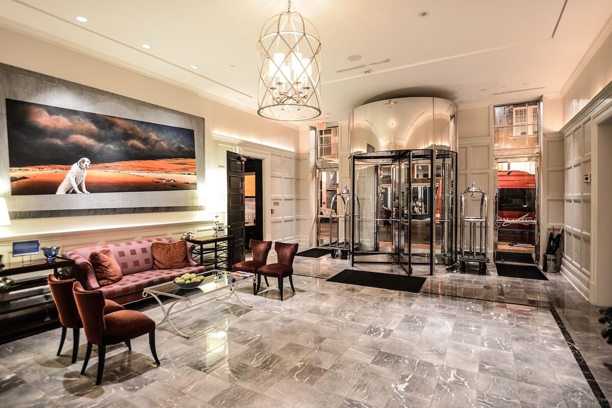 A hotel lobby with marble floors, a glass revolving door, cozy seating, a luggage cart, and a large painting of a dog welcomes guests visiting Greenwood MS for all their travel reasons.