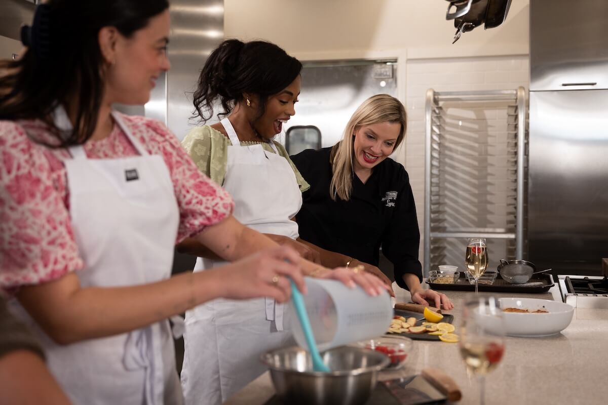 Three women in a kitchen—two in white aprons and one in black—prepare food and smile together at a counter with mixing bowls, showcasing the warmth and hospitality that are great reasons to visit Greenwood MS.