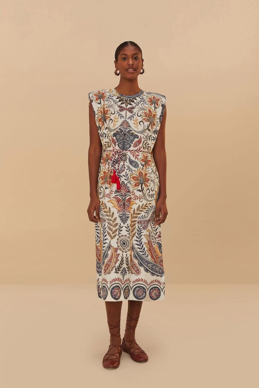 A woman stands facing forward wearing a sleeveless, knee-length dress with colorful floral and geometric patterns.