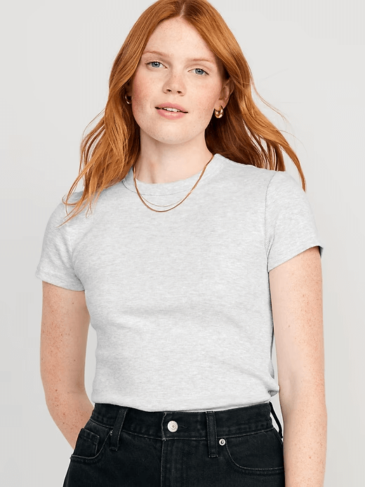 Person with long red hair wearing a light gray short-sleeve t-shirt—one of the summer essentials—black pants, gold hoop earrings, and a gold necklace, standing in front of a plain background.