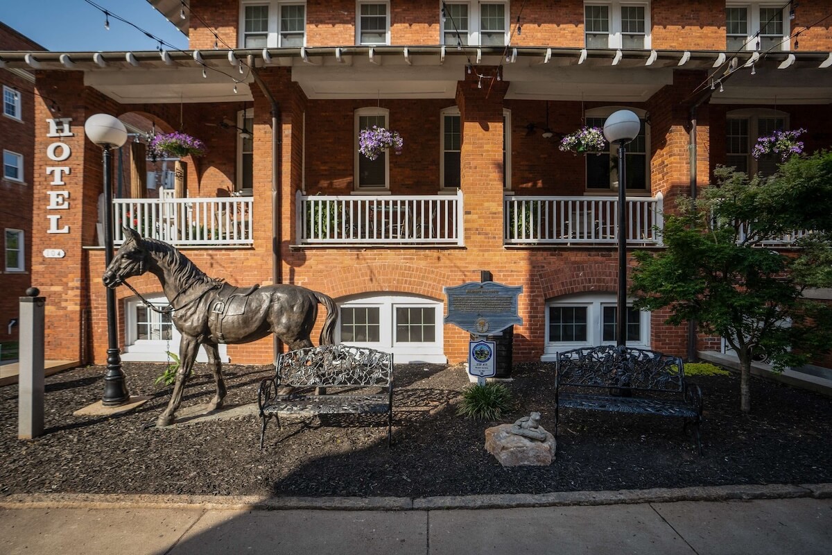 5 Charming Bed & Breakfasts in SC's Old 96 District - 4 A metal horse statue stands between two benches in front of a brick hotel building with balconies and hanging flower baskets, adding charm to one of South Carolina’s inviting Bed & Breakfasts in SC’s Old 96 District.