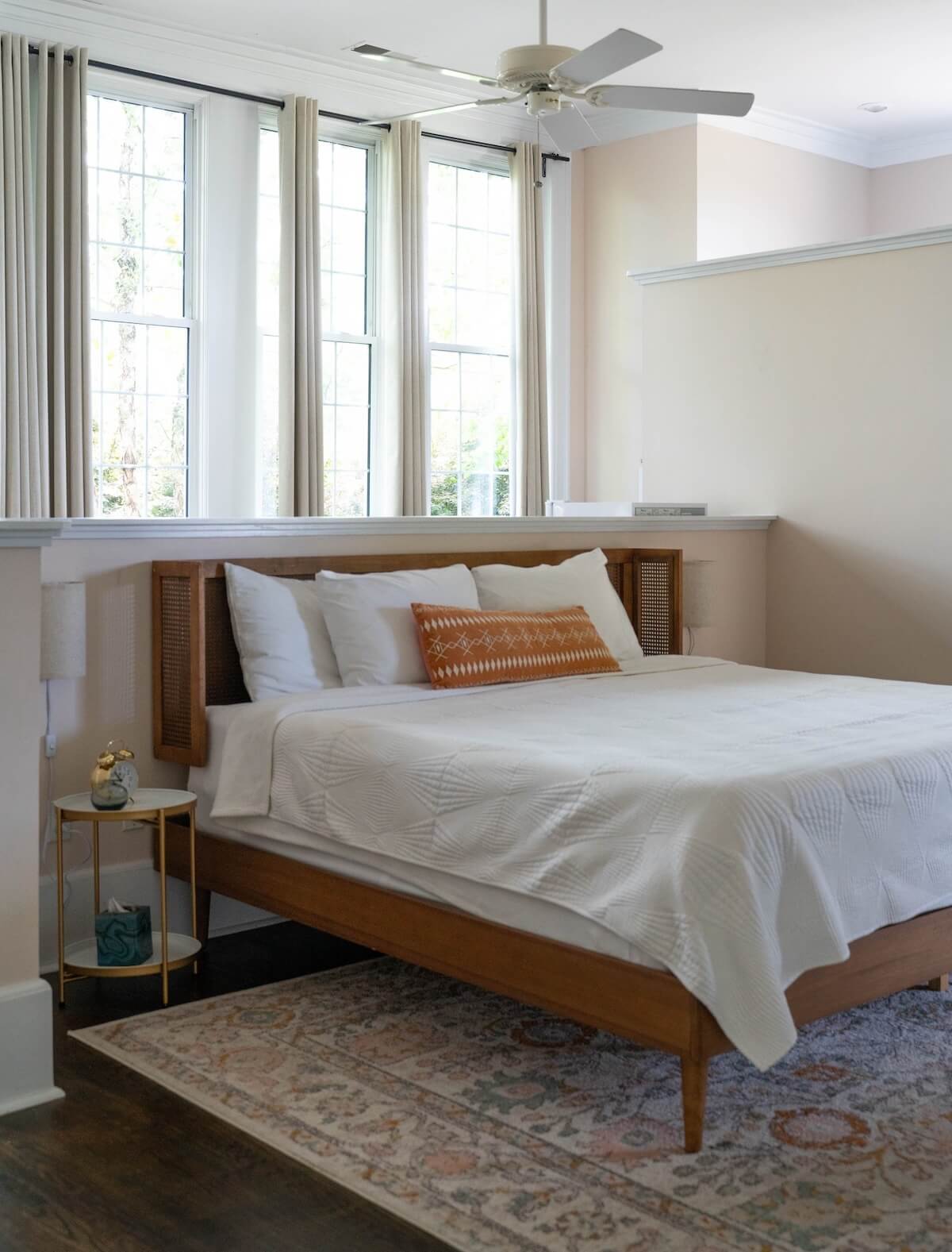 5 Charming Bed & Breakfasts in SC's Old 96 District - 3 A neatly made bed with white bedding and a single decorative pillow stands next to a small round side table in a bright room with tall windows and a ceiling fan, capturing the charm of Bed & Breakfasts in South Carolina’s SC Old 96 District.