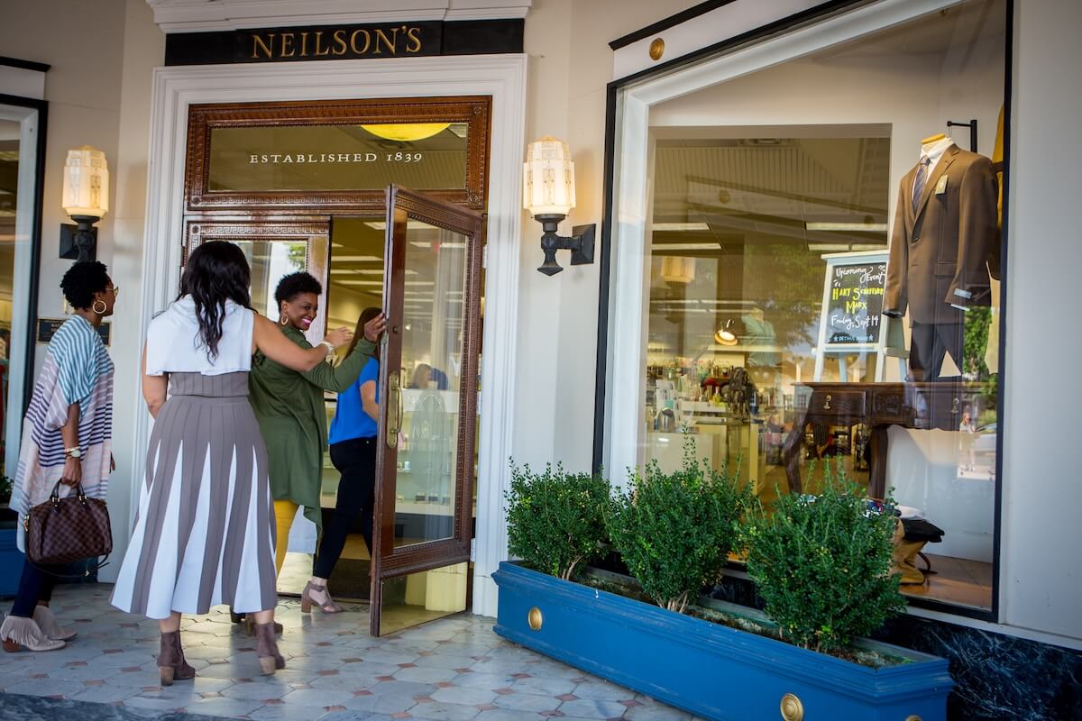 Three women enter Neilson's, a historic clothing store established in 1839, while another stands nearby with a handbag—a classic stop for any Oxford Mississippi travel or Girls’ weekend Oxford MS itinerary. A suit is displayed in the window.