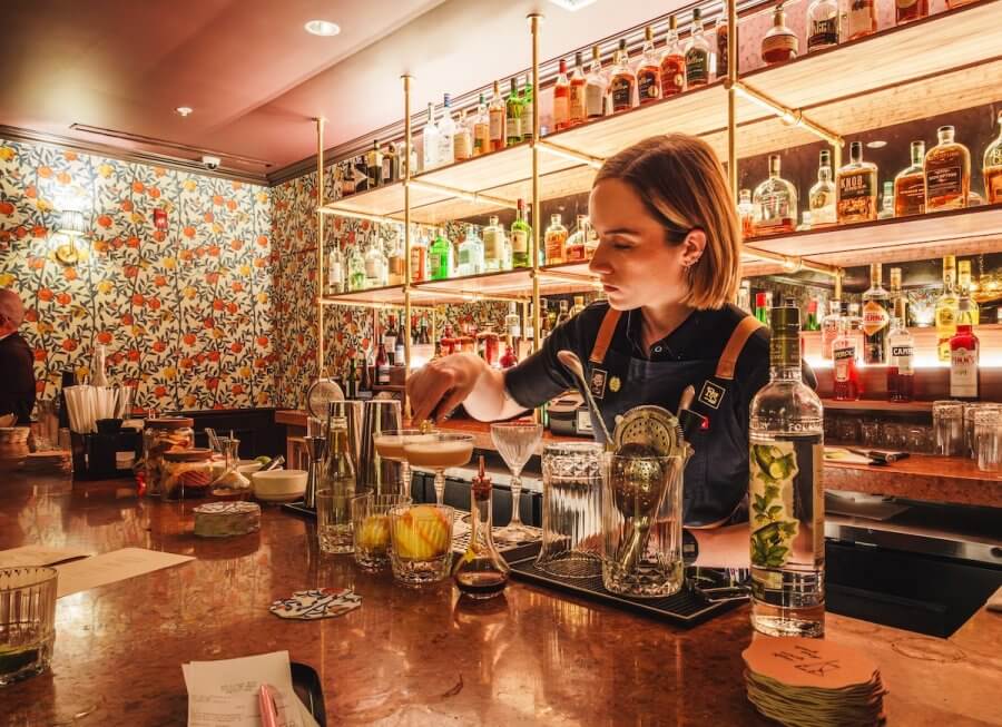 An Ode to Oxford: How to Eat Your Way Through This Delicious Small Town - 5 A bartender prepares a cocktail behind a bar lined with bottles of liquor, glassware, and fruit garnishes. Floral wallpaper adds charm in the background.