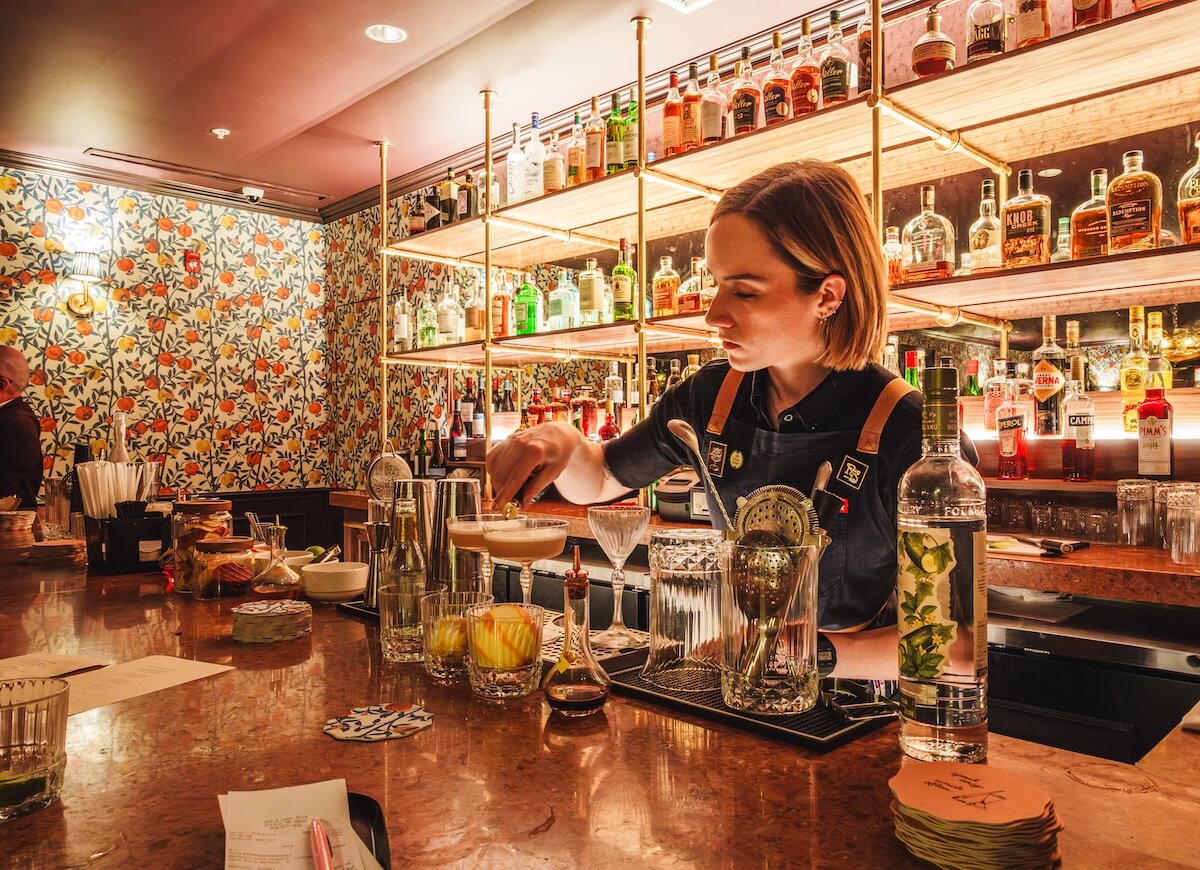 A bartender prepares a cocktail behind a bar lined with bottles of liquor, glassware, and fruit garnishes—an ideal spot to add to your Oxford MS itinerary for a memorable girls’ weekend. Floral wallpaper adds charm in the background.