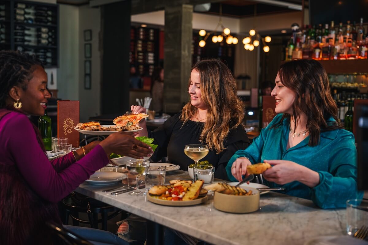 Three women enjoy a girls weekend in Oxford MS, sharing food and drinks at a restaurant table filled with pizza, salad, and appetizers.