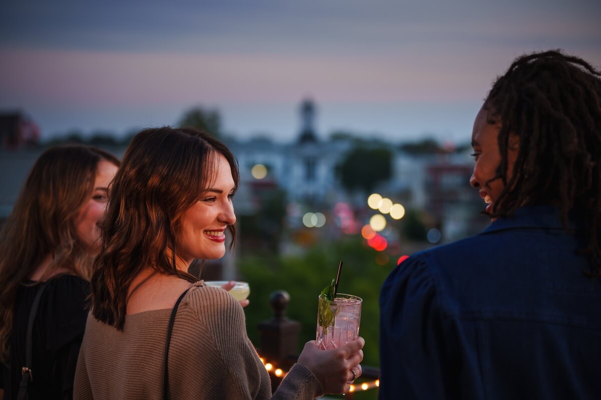 Three women stand outdoors in the evening, holding drinks and chatting—perfect for a Girls’ Weekend Oxford MS—with blurred city lights and buildings glowing behind them.
