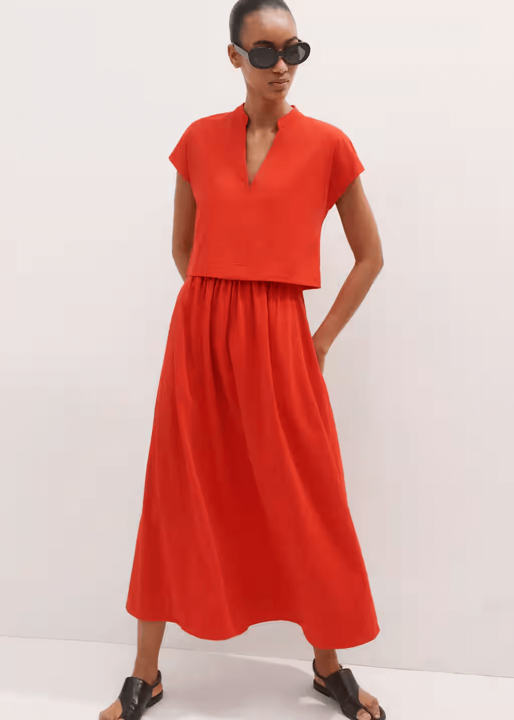 A person wearing a bright red, short-sleeve, v-neck, ankle-length dress pairs it with black sunglasses and sandals, standing against a plain light-colored background.