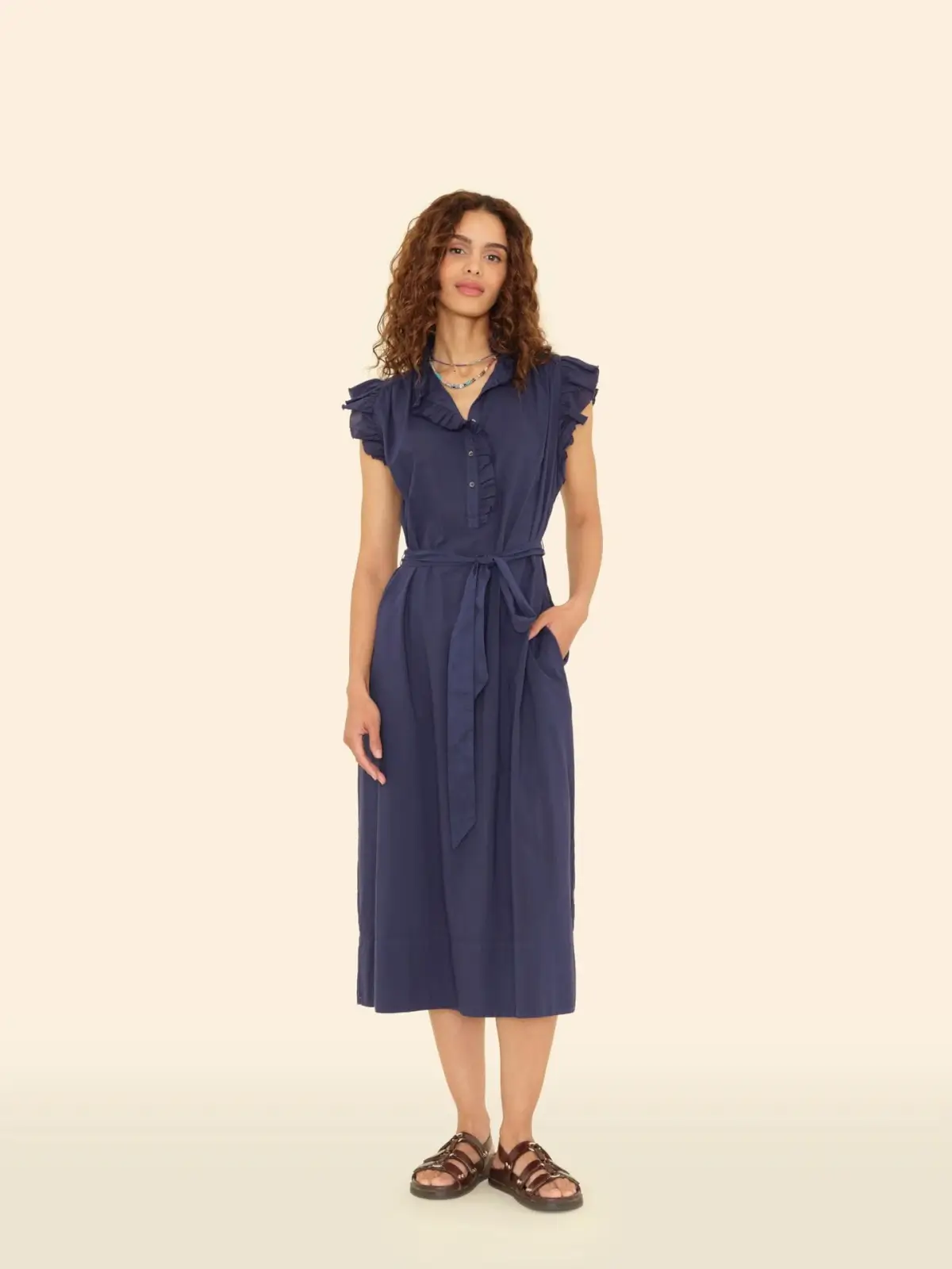 A woman stands against a plain background wearing a navy blue midi dress with ruffled sleeves, a tied waist belt, and brown sandals.