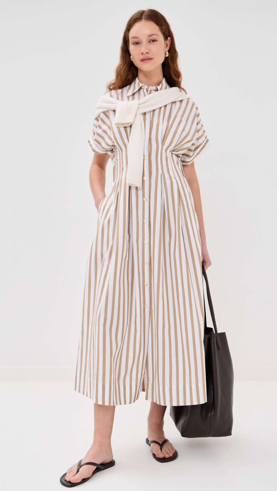 A woman wears an easy summer dress—a short-sleeve, button-down, beige and white striped midi—with a white sweater over her shoulders, black sandals, and a large black tote bag. 