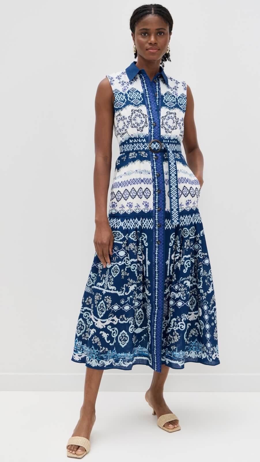 A woman stands against a plain background wearing an easy summer dress—a sleeveless, collared, button-front style with a blue and white patterned design and matching belt. She has braided hair and is wearing slide sandals.