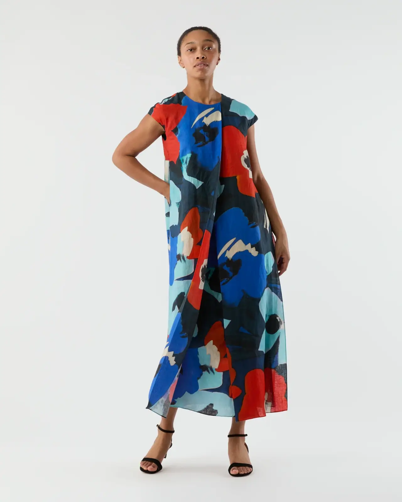 A woman stands wearing a sleeveless, ankle-length dress with a bold abstract print in red, blue, black, and teal against a plain background.