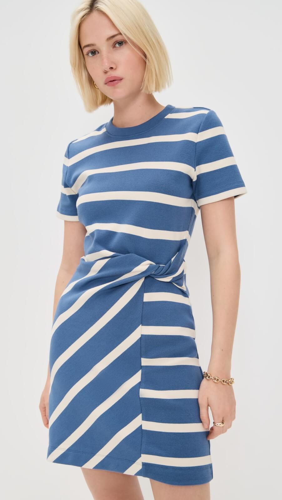 A woman wearing a short-sleeved blue and white striped dress with a knot detail at the waist stands against a plain white background, showcasing one of this season’s easy dresses.