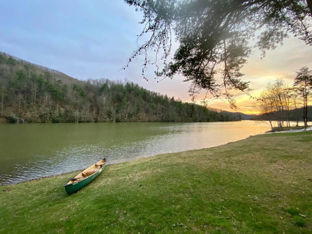 A green canoe rests on a grassy shore beside a calm lake, surrounded by trees, with the sun setting in the background—a must-visit Virginia scene for anyone seeking serene and unforgettable Virginia travel experiences.