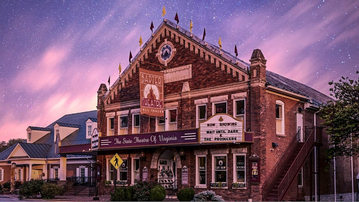 Historic brick building with a marquee reading "Barter Theatre," decorative trim, arched windows, and a sign listing current shows—this must-visit Virginia destination shines under a twilight sky with clouds and stars.