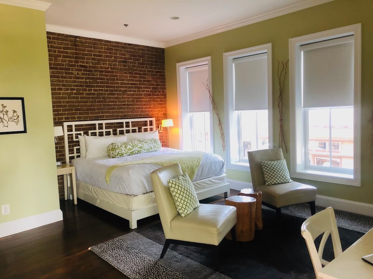 A bedroom in Virginia with a brick accent wall, a bed with white bedding, two cream chairs, small wooden tables, and three large windows with shades—perfect for travel lovers seeking must-visit destinations.