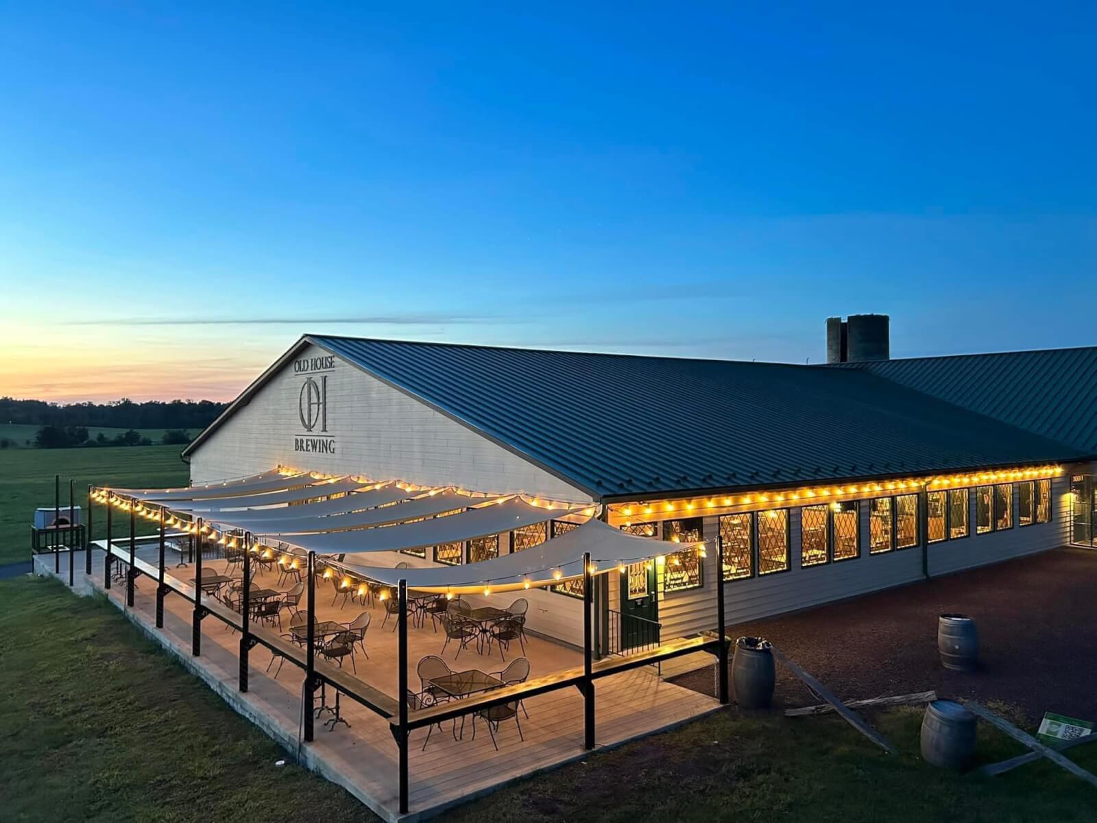 A must-visit place for travel enthusiasts, this brewery building features outdoor patio seating under string lights and canopies at dusk, set amidst open fields—one of the top Virginia destinations for a relaxing evening.