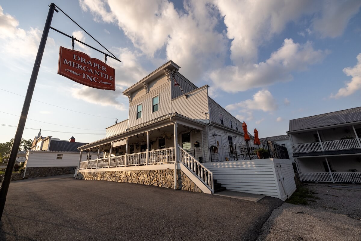 A white, two-story building with wraparound porch and a sign reading "Draper Mercantile Inc."—a charming stop among must-visit places for those looking to travel Virginia under a partly cloudy sky.