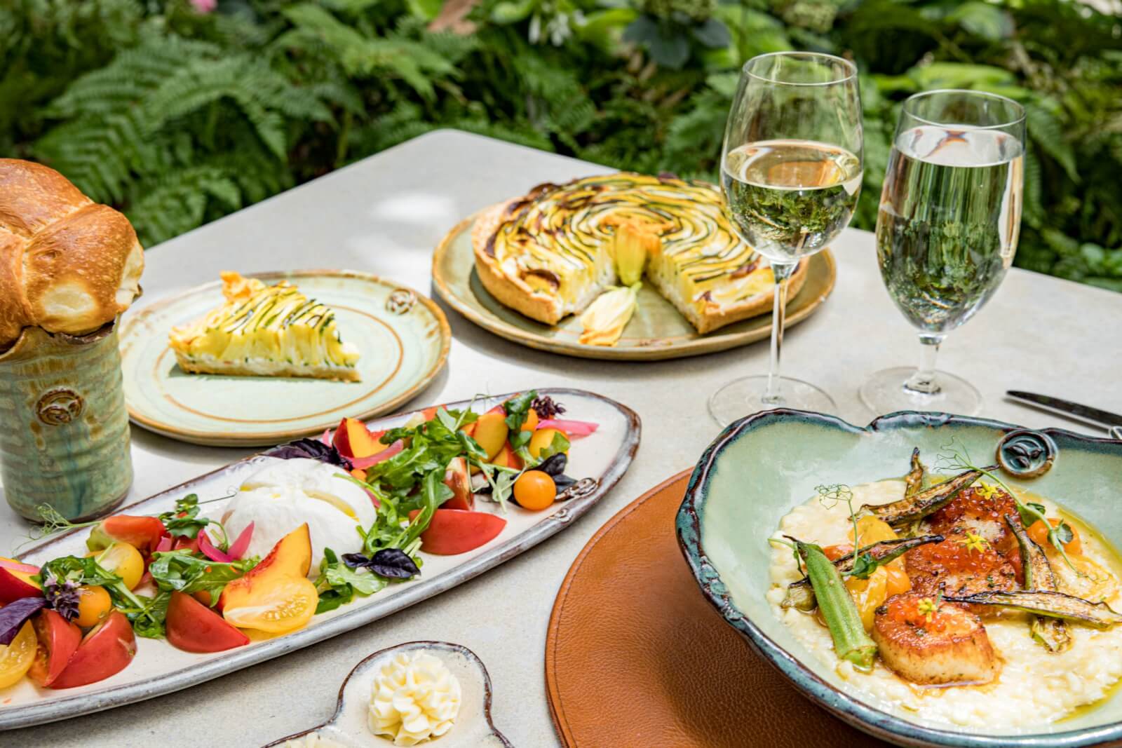 A table set outdoors among greenery features a savory galette, salad with peaches and burrata, fish with vegetables on risotto, popover bread, and two glasses of white wine.