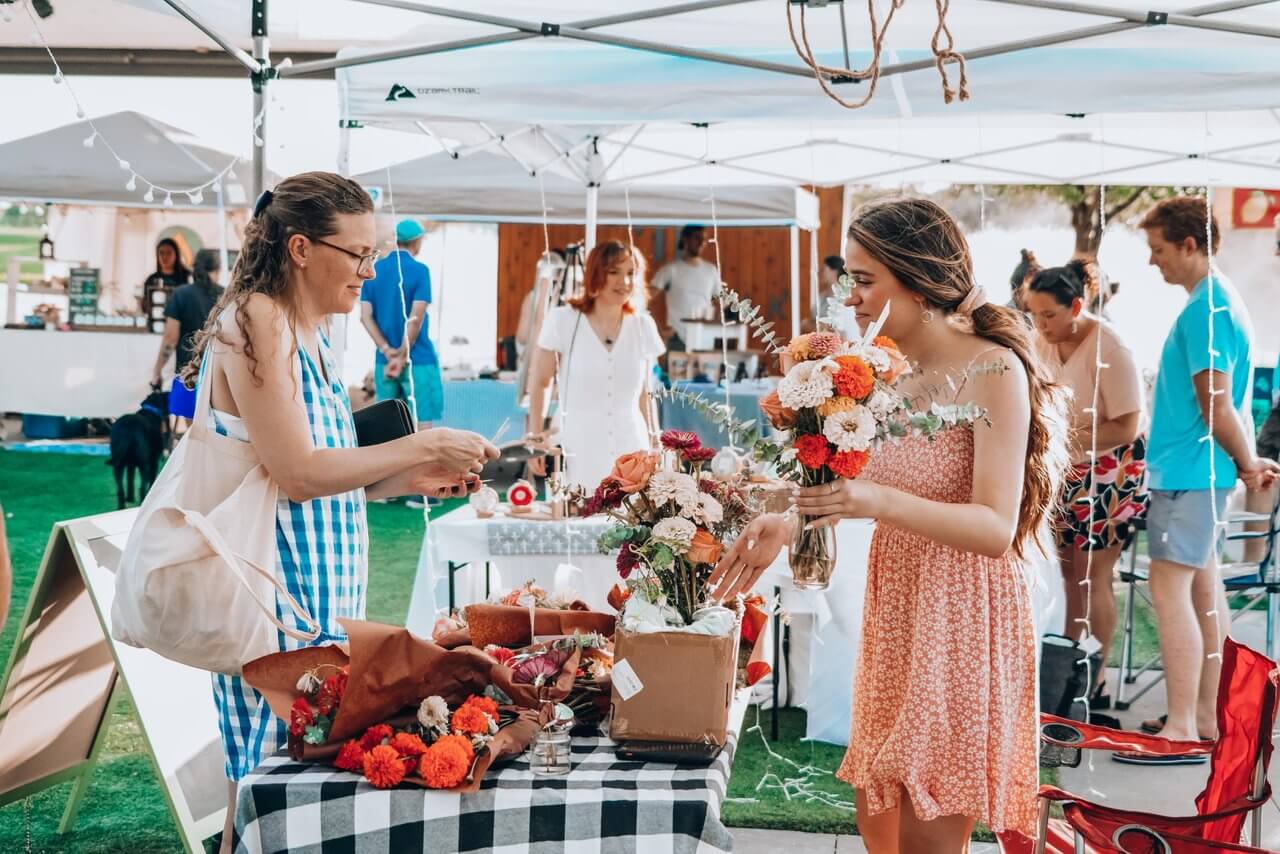 Two women interact at a market stall; one holds a bouquet of flowers while the other pays. Other people and booths bustle in the background.
