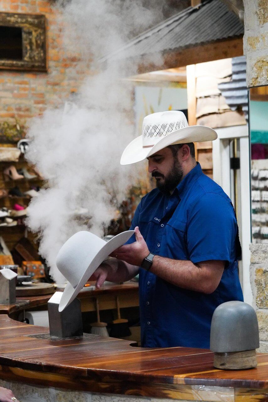 A man in a blue shirt shapes a white cowboy hat using steam at a wooden counter in a hat shop, capturing the classic style.