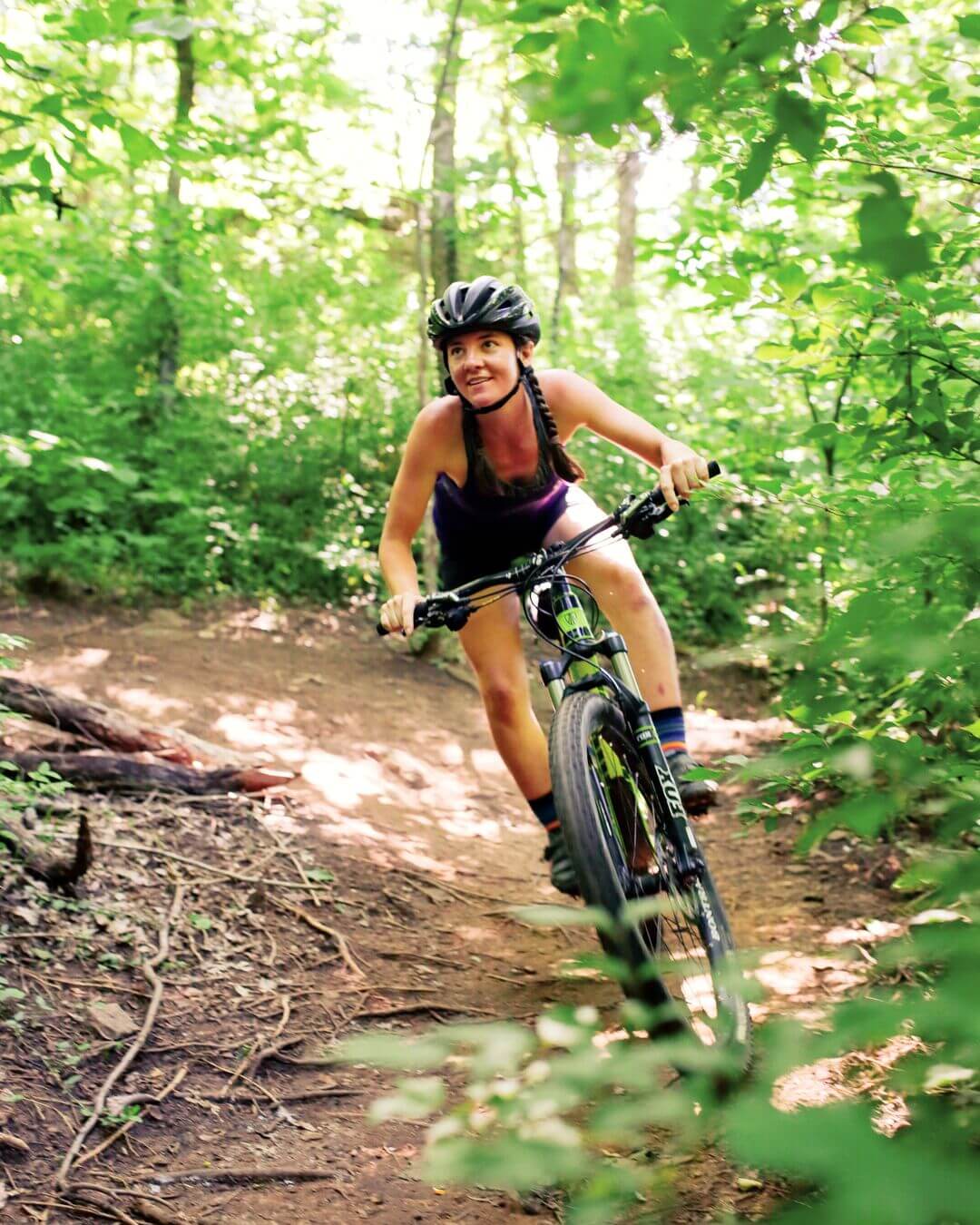 A woman wearing a helmet rides a mountain bike on a dirt trail through a lush, green forest, reminiscent of the scenic landscapes found near SEC Towns.