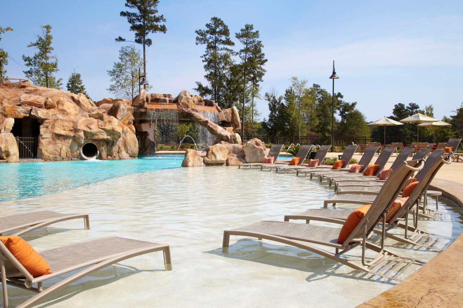 Chairs with orange cushions line a shallow pool area near a rock formation with a water slide and waterfall, creating a relaxing retreat, surrounded by trees and umbrellas.