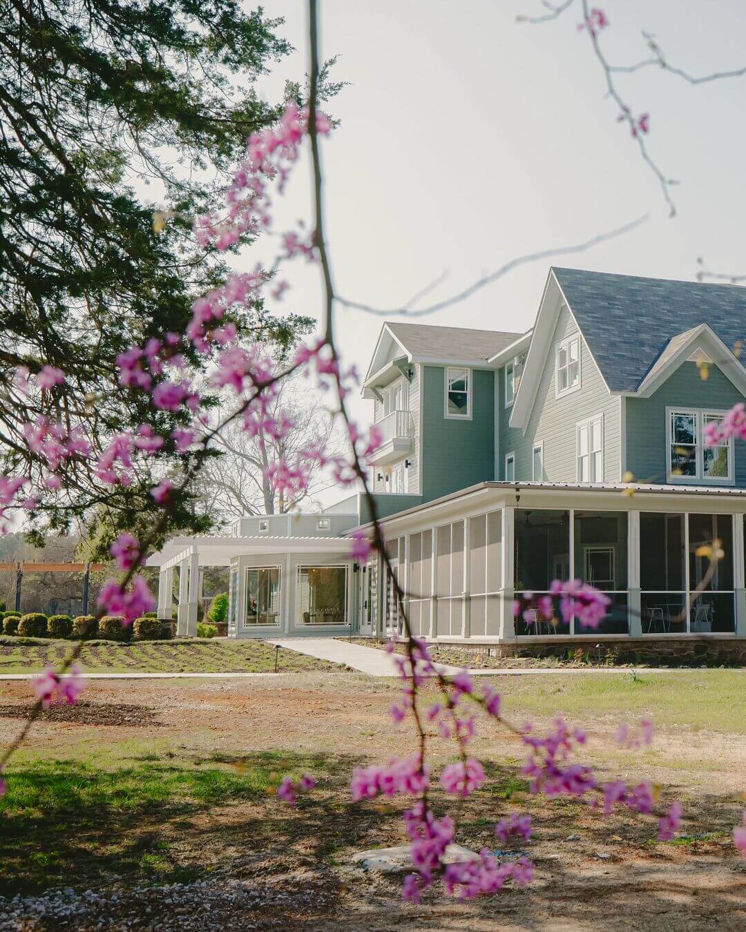Light green two-story house with a screened porch, seen through branches with pink flowers on a sunny day—classic charm reminiscent of SEC Towns.